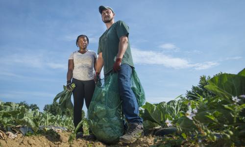 Two farmers stand side by side, both holding crops