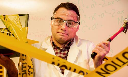 A confused scientist holds up a hammer and broken laptop