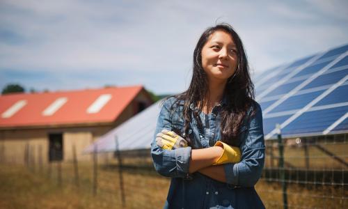 A person stands in front of a solar panel with their arms crossed