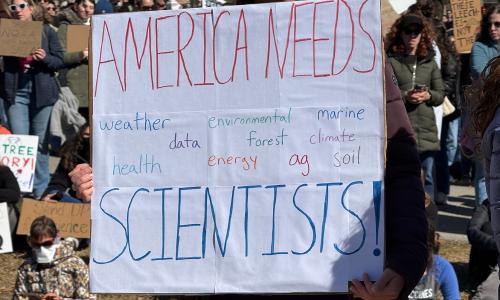 A person holding a sign that reads "AMERICA NEEDS SCIENTISTS!"
